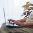 Older adult sitting by a window using a laptop computer, researching online with natural light and a houseplant in the background.