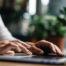 Close-up of a person typing on a laptop keyboard, working at a desk with green plants blurred in the background — representing focus, productivity, or completing an online task.