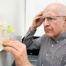 An older man wearing glasses and a checkered shirt looks intently at a handwritten note taped to a wall or refrigerator, appearing focused and slightly puzzled as he reads reminders surrounded by colorful sticky notes — illustrating memory difficulties or forgetfulness.