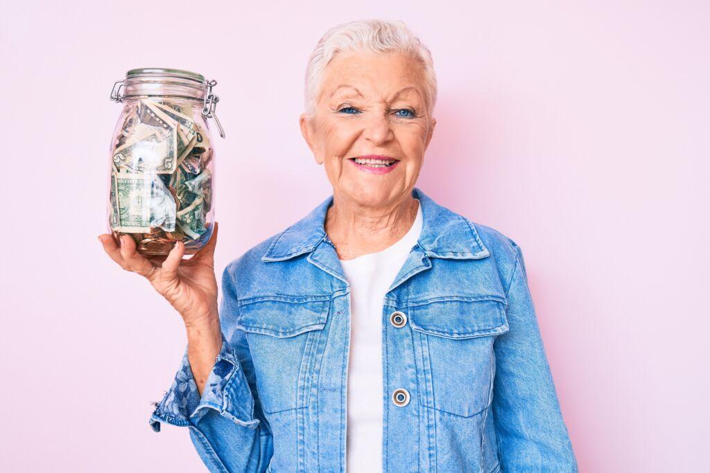 Smiling older woman holding a large jar filled with cash against a pink background.