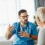 A healthcare professional wearing blue scrubs and a stethoscope speaks with an older male patient in a bright medical setting, explaining something with hand gestures — representing a doctor–patient consultation or discussion about health concerns.