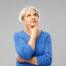 Older woman with short gray hair wearing a blue sweater, looking up thoughtfully with her hand on her chin against a plain light gray background