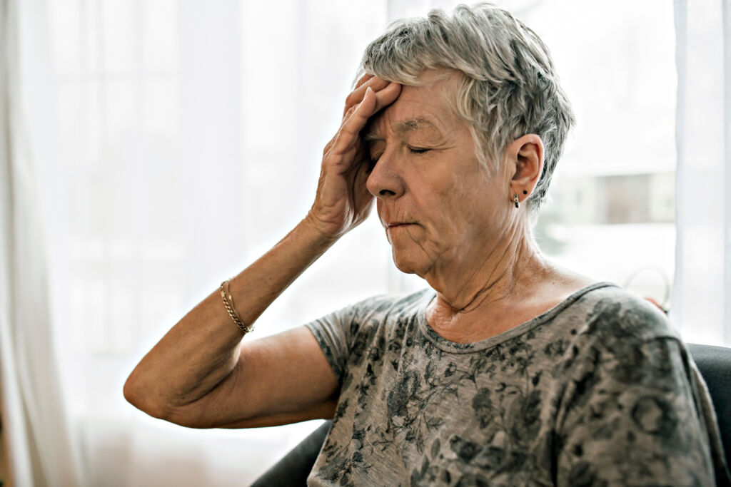 Older woman sitting indoors with her hand on her forehead, appearing thoughtful or concerned.